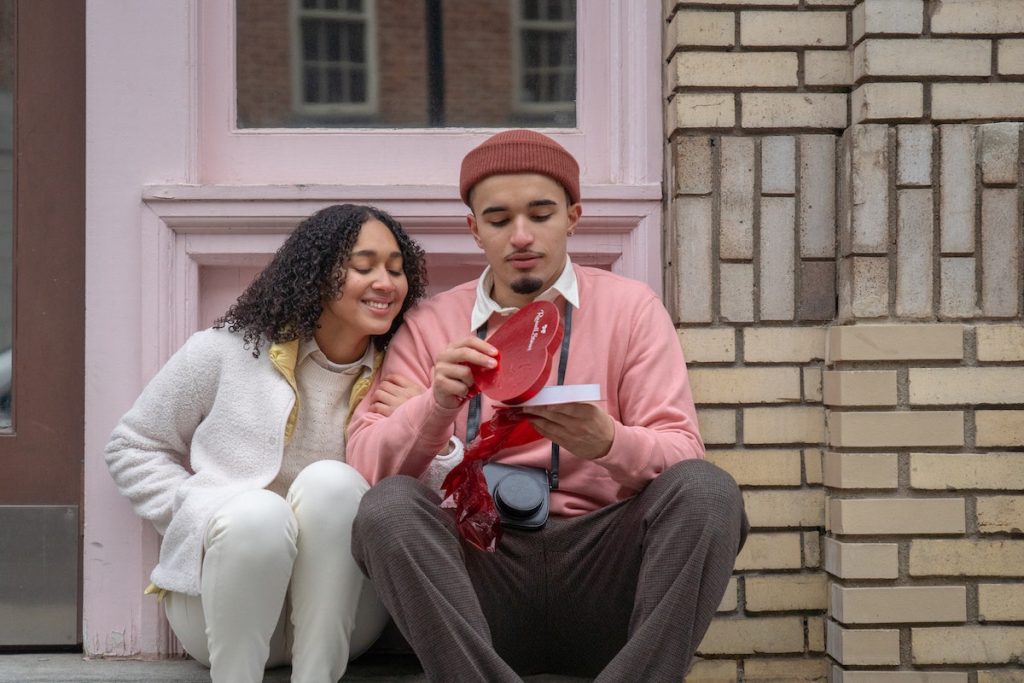 Young Hispanic man opening gift box sitting on street near girlfriend with closed eyes